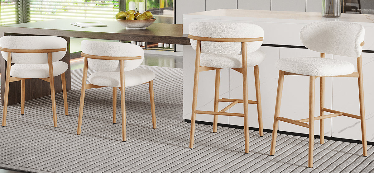 Contemporary kitchen/dining area with wood-top table, white marble island, light wood chairs, and stools with plush white upholstery. Pears in a bowl and a notebook rest on the table.