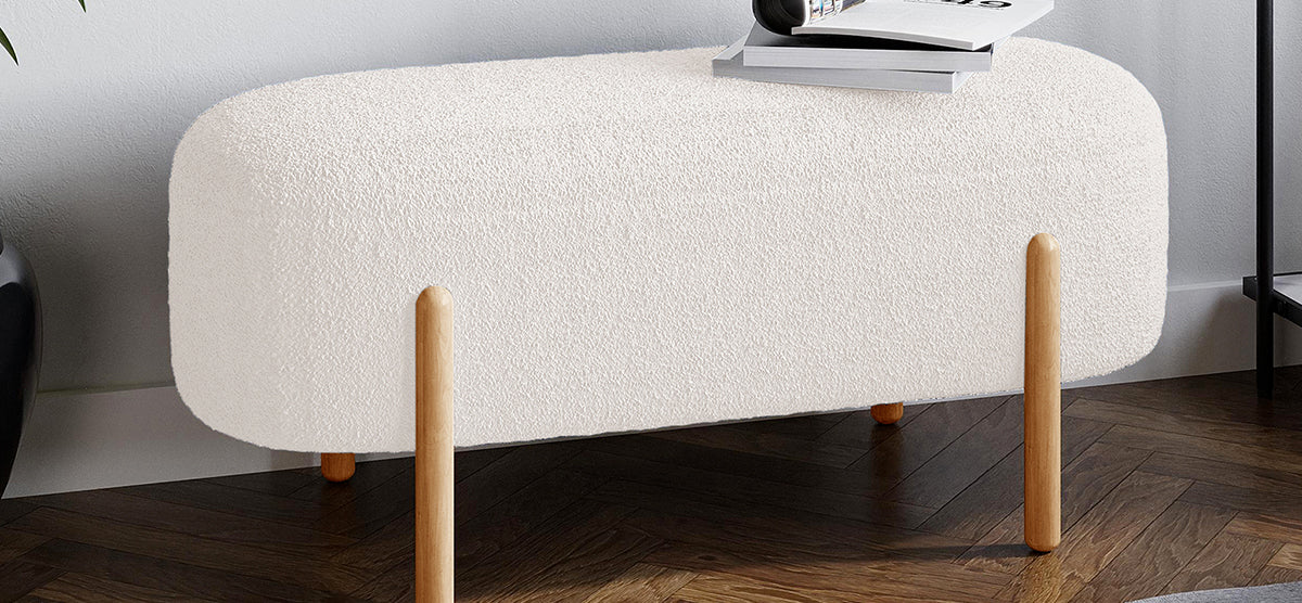 Small rectangular ottoman with rounded corners, upholstered in off-white bouclé fabric, on four light wood legs. Topped with stacked magazines, set on dark herringbone-patterned wood floor.