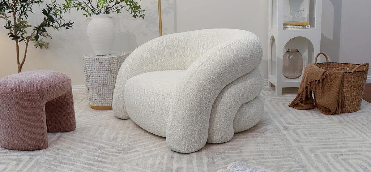 A modern lounge corner featuring a creamy white boucle armchair, mauve-pink pouf, stone-mosaic side table with vase, olive tree, white shelving, wicker basket, and geometric rug.