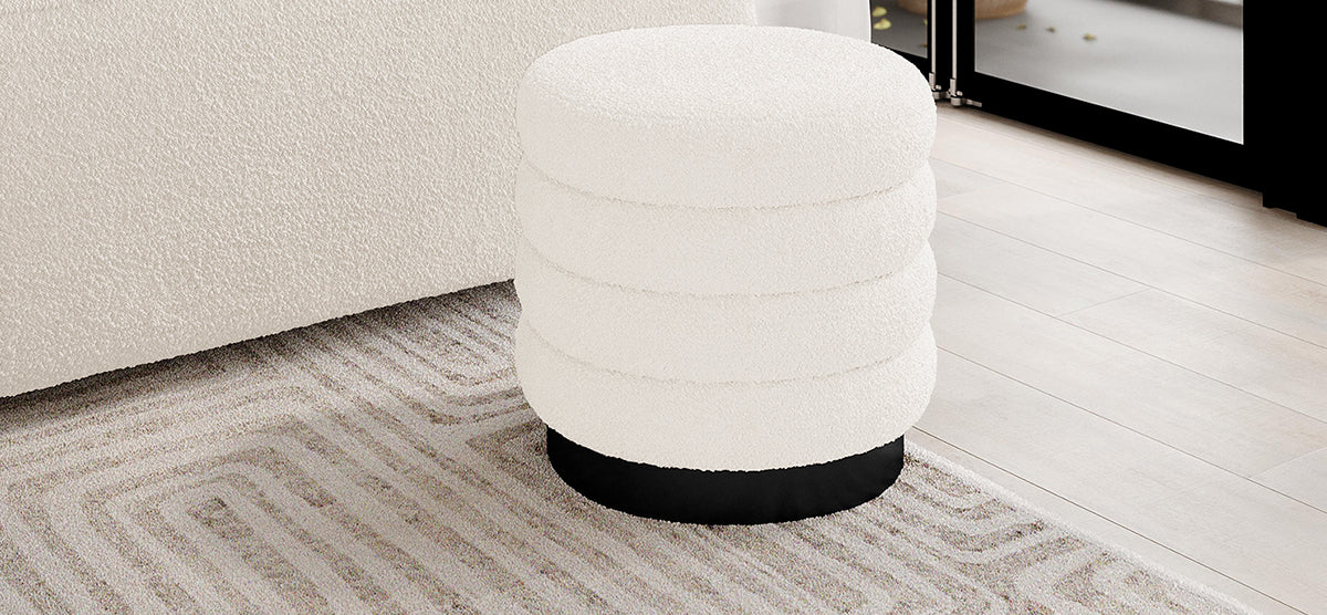 Cream-white bouclé upholstered ottoman with four padded rings and a black base on a patterned rug. Positioned beside a light sofa near a glass door, creating a cozy lounge setting.