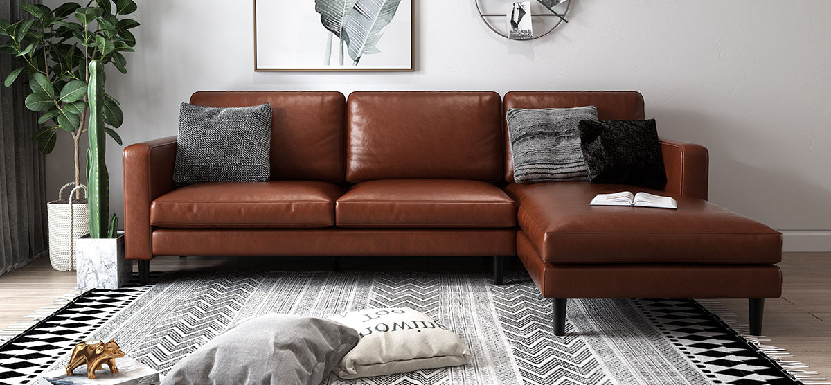 Modern living room with a cognac-brown leather sectional sofa, gray throw pillows, geometric rug, potted plants, botanical print, wall mirror, floor cushions, and a gold bull figurine.