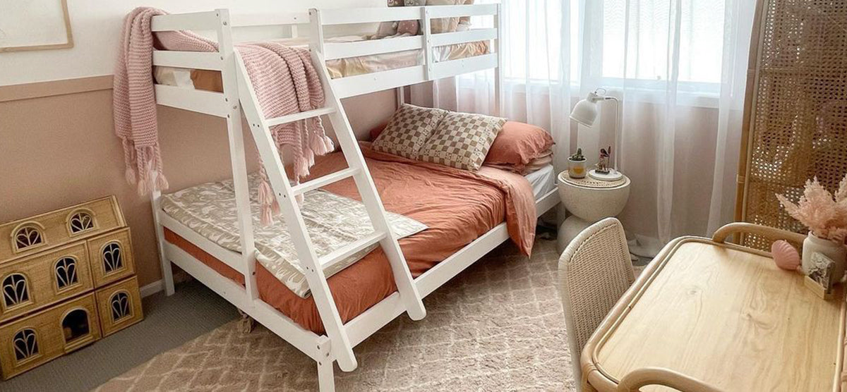Pastel-toned bedroom featuring a white bunk bed with blush and terracotta bedding, a wooden dollhouse, a round side table with decor, and a woven chair.