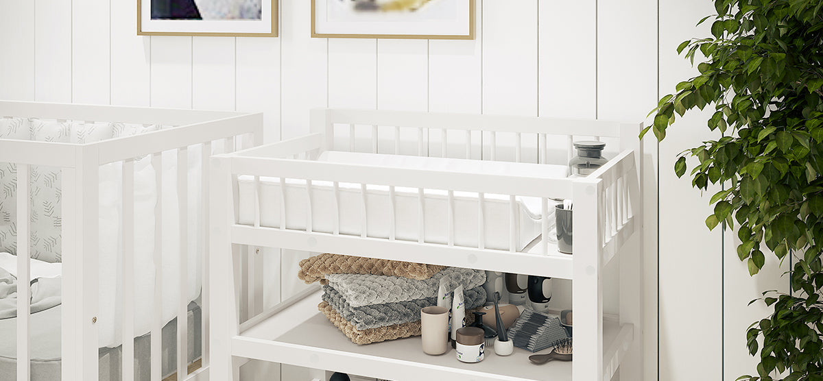 Minimalist nursery corner with white wooden crib and matching changing table. Neutral-tone blankets, baby-care items on shelf. Shiplap wall with art prints and potted houseplant beside.