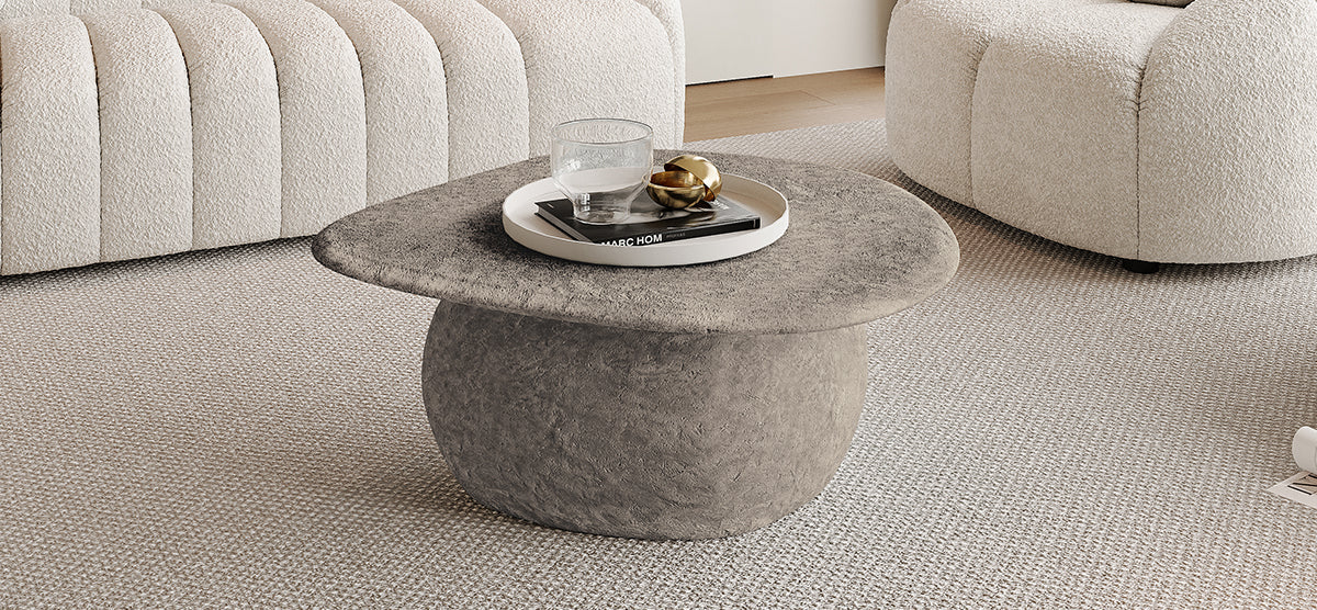 Minimalist living area with a stone-gray round coffee table featuring a white tray, tumbler glass, books, and gold decor. Background shows off-white bouclé sofas on beige carpet.