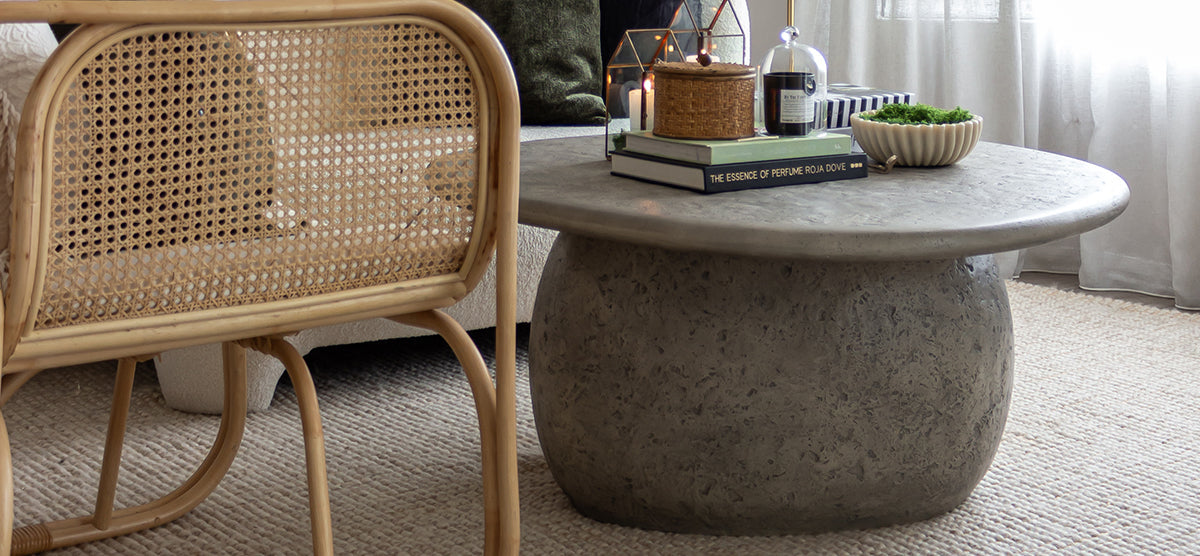 Cozy living-room scene with a round concrete coffee table displaying books, candles, a wicker box, a plant bowl, and adjacent rattan accent chair on a textured rug.