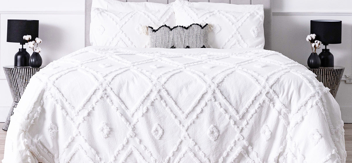 Minimalist bedroom featuring a white textured duvet with diamond-and-dot pattern, black and white accent pillow, black ribbed side tables, and black table lamps with vases.