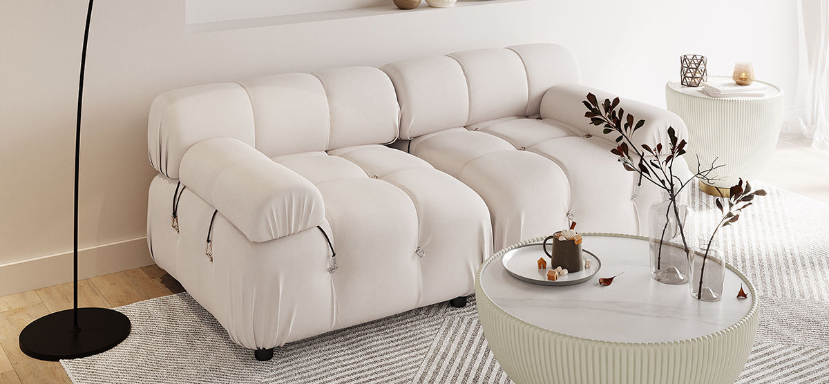 A modern living area featuring a tufted cream-colored sofa, round coffee table with vases and a mug, black floor lamp, and geometric-patterned rug on wood flooring.