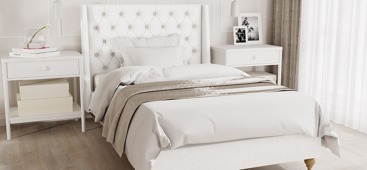 Minimalist bedroom with a white upholstered bed featuring a tufted headboard. Flanked by white nightstands; left with books and vase, right with framed art. Light wood floor, sheer curtains.