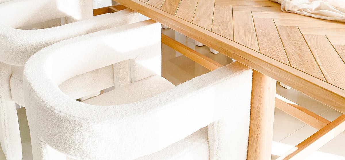 Solid-wood dining table with herringbone plank top, surrounded by plush, white boucle-upholstered armchairs, illuminated by natural light highlighting the warm wood tones and soft textures.