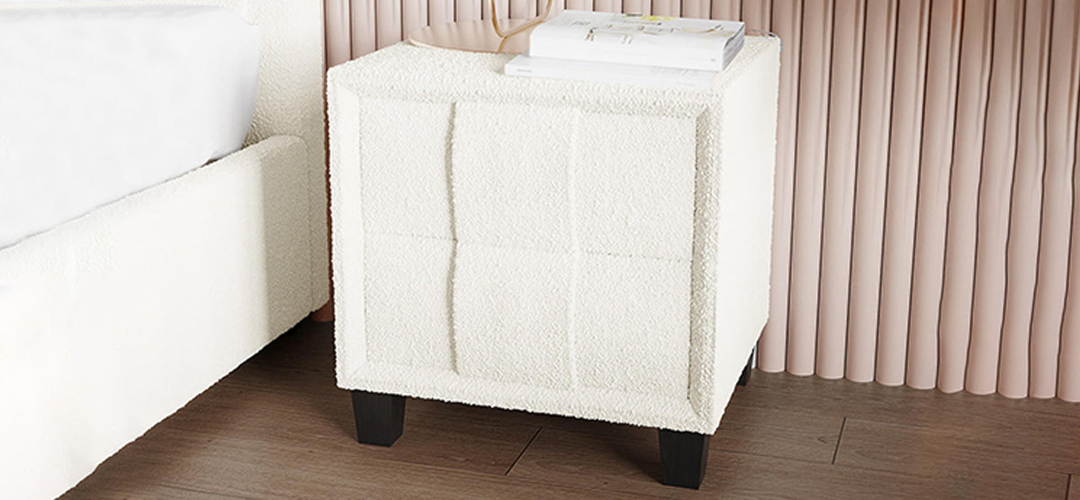 Cube-shaped upholstered ottoman in textured off-white fabric, with four dark wooden legs, next to a bed. Topped with a book, possibly a lamp, against blush drapes.