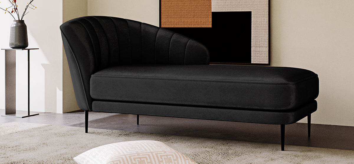 Minimalist corner with a black chaise lounge on metal legs, T-shaped side table with vase, abstract wall art, and a glimpse of a throw pillow on a neutral rug.