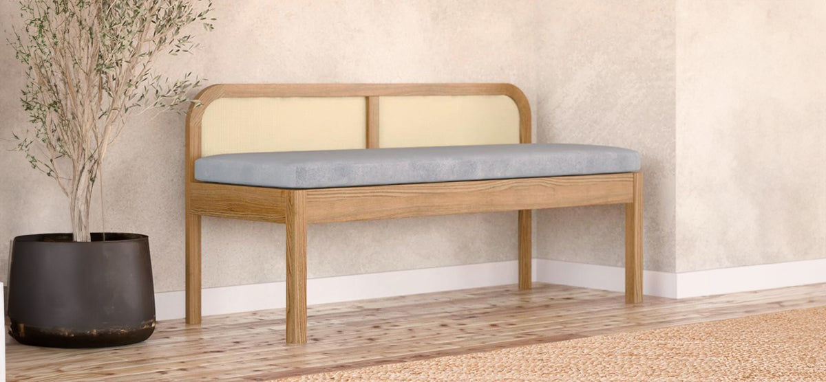 A modern entryway features a wooden-framed bench with a light-gray cushion and cane-style backrest, accompanied by a slender plant in a matte pot on hardwood flooring.