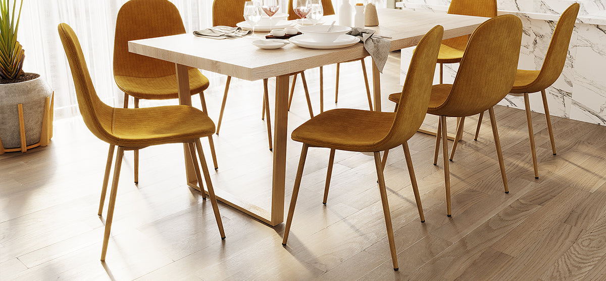 A modern dining area with a wooden table set for six, featuring matching chairs with metal legs. Sunlight filters through sheer curtains, highlighting a potted plant and marble-accented wall.
