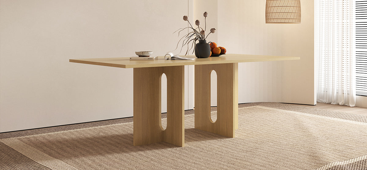 A minimalist interior with a light-wood table featuring a ceramic bowl on a book, an open magazine, a black vase with dried stems, and oranges in a dish.