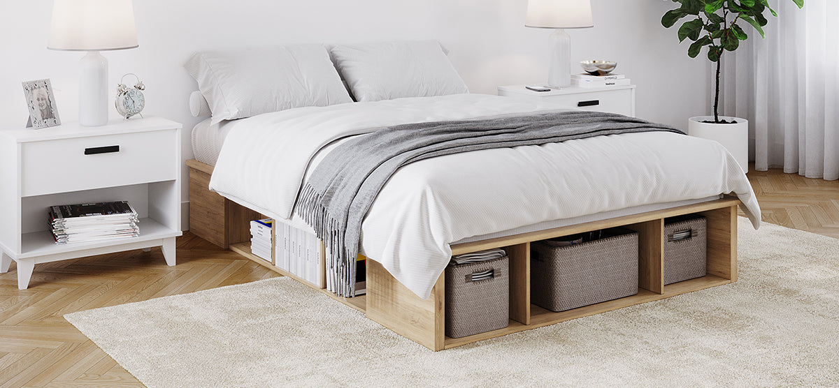 A minimalist bedroom with a light-wood platform bed featuring cubby storage, white linens, matching nightstands, a herringbone floor, a plush rug, and a potted plant by sheer curtains.
