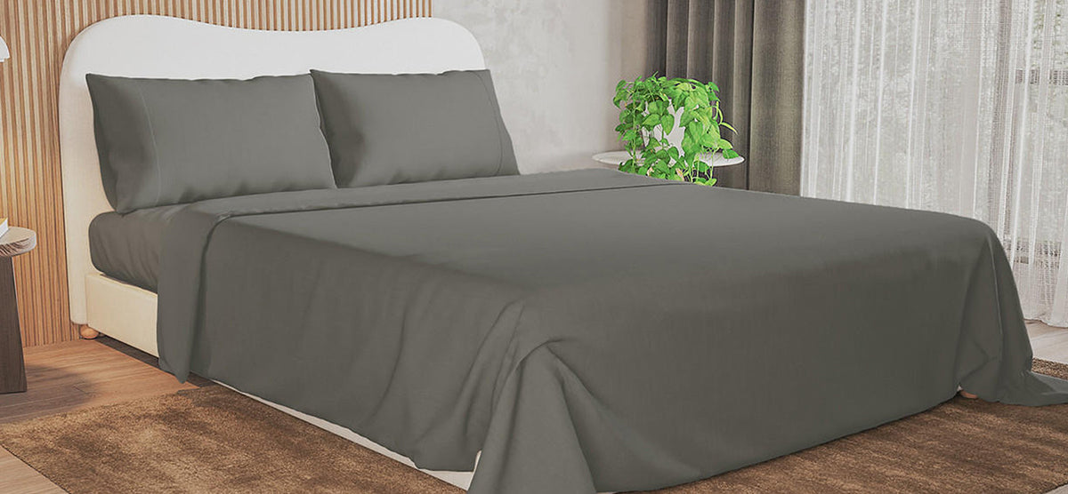 Double bed with white upholstered headboard, dark gray bedding, and gray pillows. Accompanied by a wooden nightstand with a lamp and a side table with a potted plant.