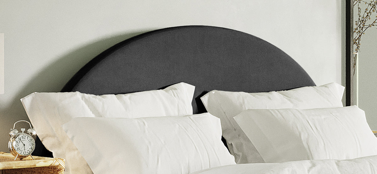 A neatly made bed with a dark upholstered headboard and four white pillows, flanked by a wicker side table with a silver twin-bell alarm clock.
