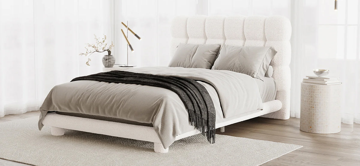Minimalist bedroom with a white boucle-upholstered platform bed, tufted headboard, gray bedding, knit throw, side table with books, lamp, and vase with branches, light wood floor.