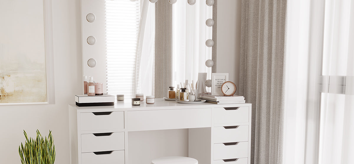 A minimalist dressing area with a white vanity featuring a “Hollywood”-style mirror, organized cosmetics, books, a clock, a white stool, and a nearby snake plant.