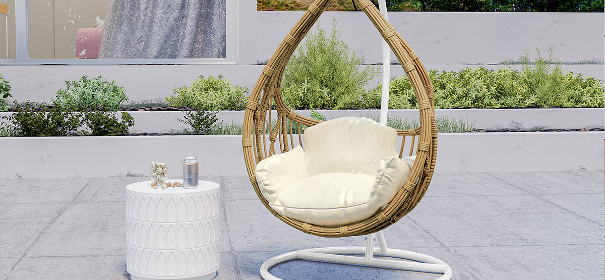 Modern outdoor nook featuring a woven rattan hanging egg chair with ivory cushion, accompanied by a white geometric side table holding a drink can and decorative shells.