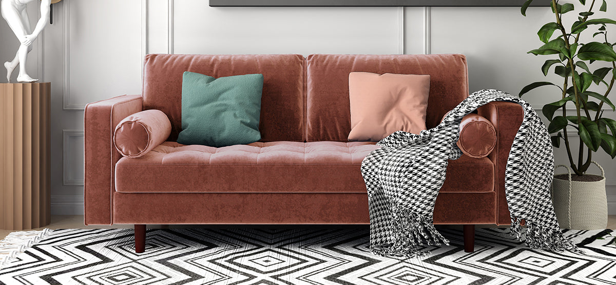 Rust-colored velvet sofa with bolster and accent pillows, houndstooth throw, geometric rug, fluted side table with sculpture, leafy plant, and framed artwork above.