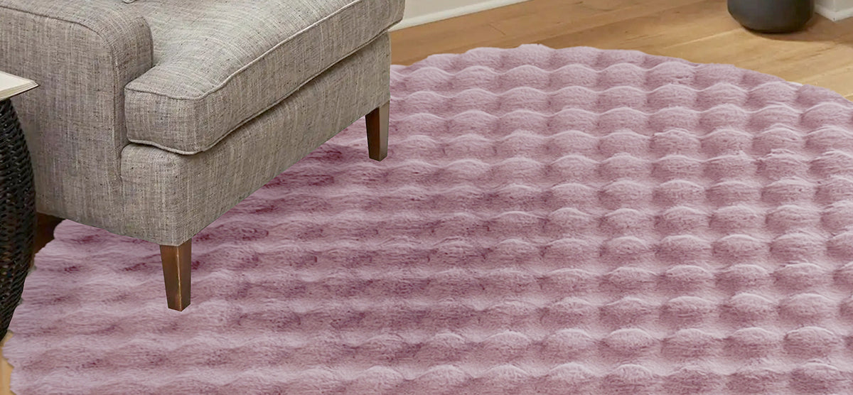 Gray upholstered sofa corner with a wooden leg, placed on a round, bubble-textured pink faux-fur area rug over a light hardwood floor.