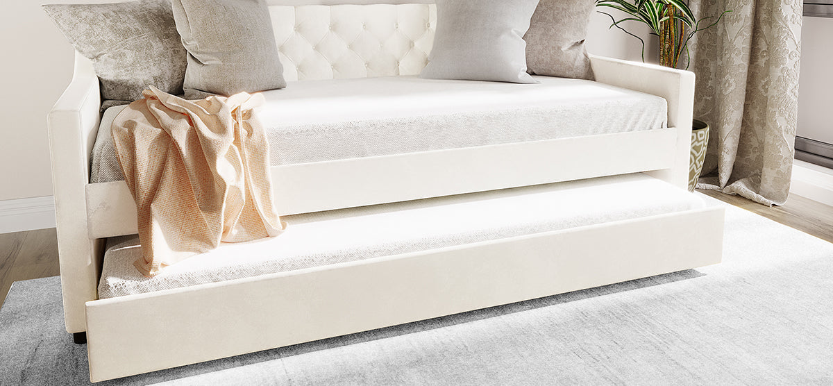 Light-colored upholstered daybed with tufted backrest, neutral throw pillows, and pull-out trundle, accompanied by a soft throw, on a rug over a wood floor.