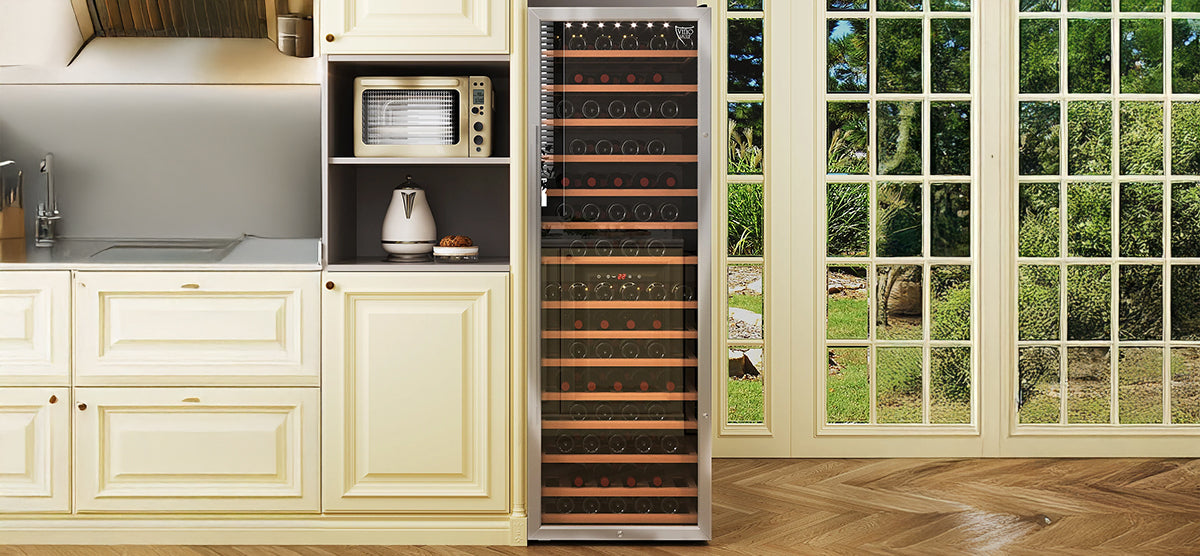 Upscale kitchen with cream cabinetry, stainless sink, hood vent, and inset shelf with microwave and kettle. Centered is a glass-front wine refrigerator. French doors overlook a garden.