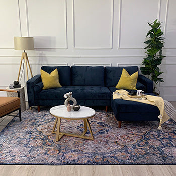 Styled living-room vignette featuring a navy-blue sectional with mustard pillows, yellow-striped throw, marble coffee table, tan leather armchair, tripod lamp, and potted plant.