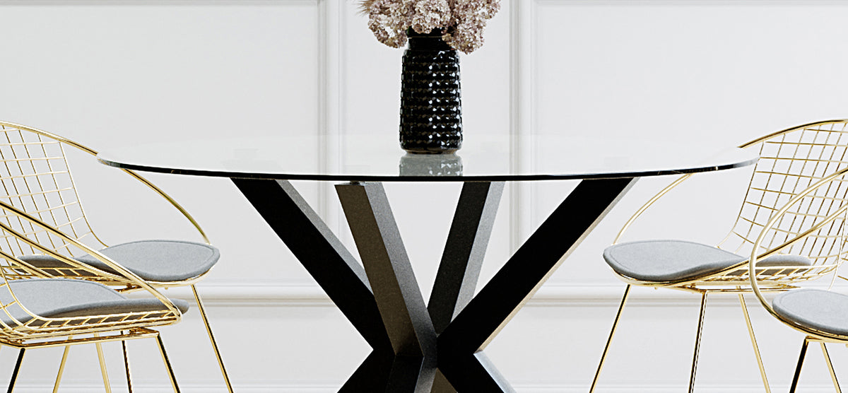 Modern dining setup with a round glass-top table featuring a black metal base. Adorned with a black vase and dried blooms, flanked by brass wire-frame chairs with gray cushions.