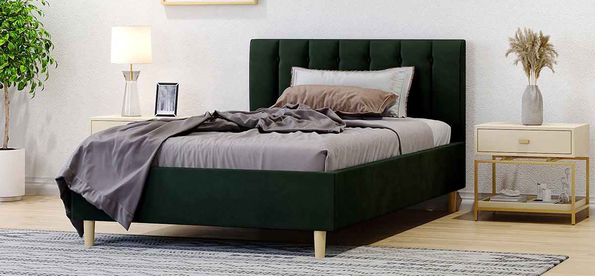 Bedroom corner featuring a deep green velvet platform bed with tufted headboard, neutral linens, wooden nightstands with decor, a leafy plant, and patterned rug on light hardwood floor.