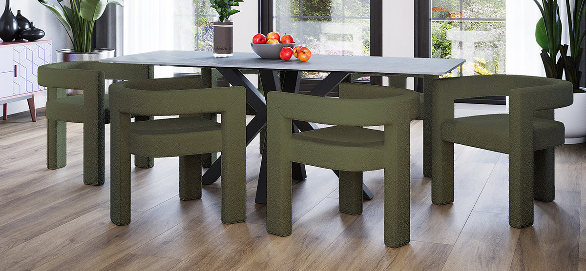 Modern dining area with a long, dark tabletop, six olive-green chairs, and a bowl of apples. Floor-to-ceiling windows reveal greenery outside. Light wood laminate flooring.
