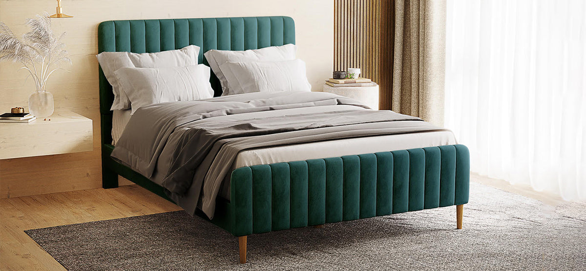 Modern bedroom featuring a teal-green velvet, channel-tufted bed with light gray bedding, flanked by a wooden nightstand and round table, both adorned with decor and books.
