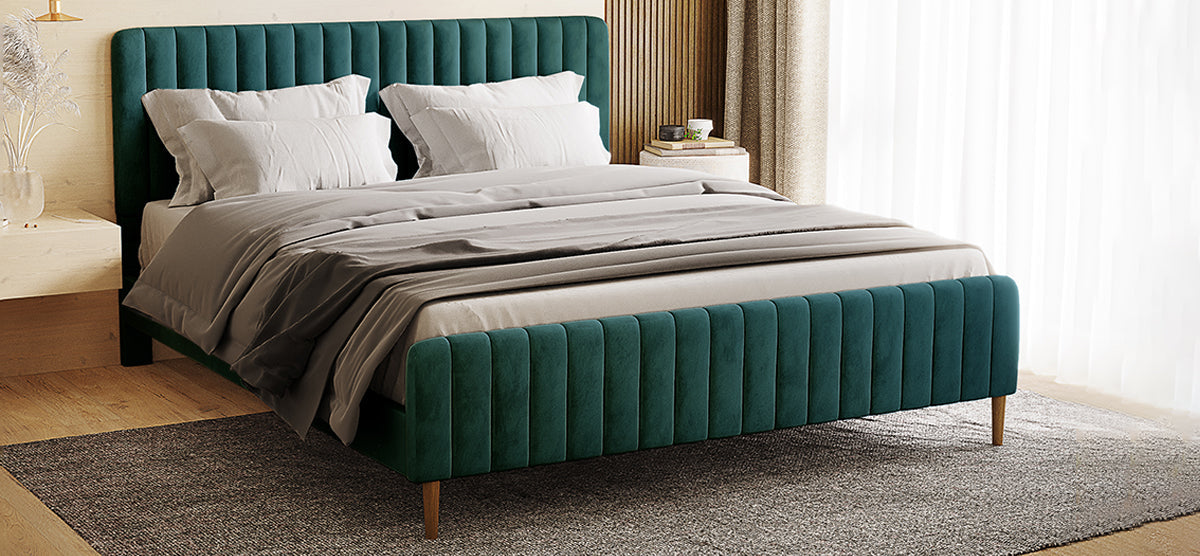 Modern bedroom with an emerald-green velvet upholstered platform bed, light gray bedding, wooden nightstands, pampas grass vase, books with mug, and sheer curtains filtering natural light.