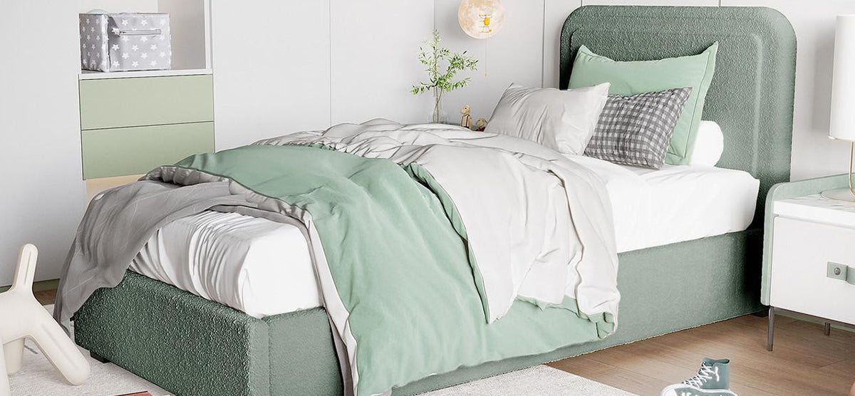 Modern bedroom with a muted sage-green upholstered platform bed, white and green bedding, white bedside cabinet with lamp, light wood floor, and minimalist decor including plants and figurines.
