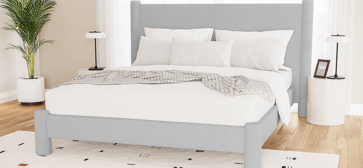 Modern bedroom with a light gray upholstered platform bed, white pillows, and a gray-and-white knit throw. Features a white side table, tall potted plant, and sheer curtains.