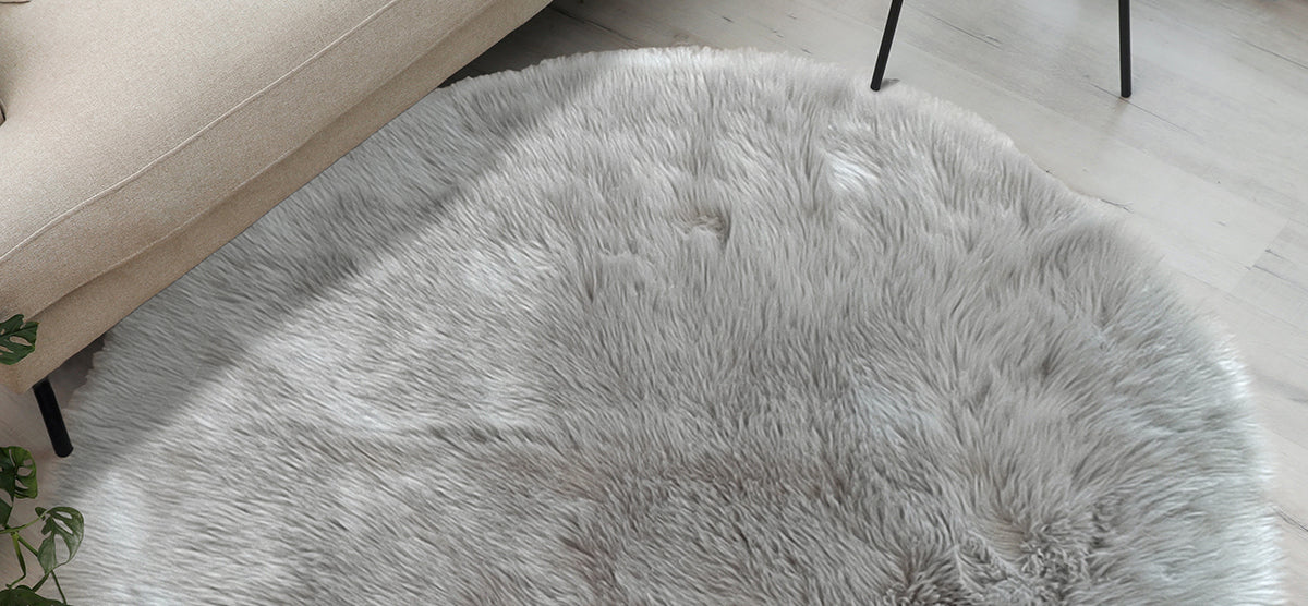 A round, shaggy gray rug under a beige upholstered sofa, with a small table leg and green plant leaves visible at the edges in a light-colored living room.