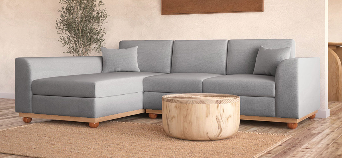 A modern living room featuring a light-gray L-shaped sofa with matching throw pillows, a round wooden coffee table, a woven area rug, and a tall potted plant.