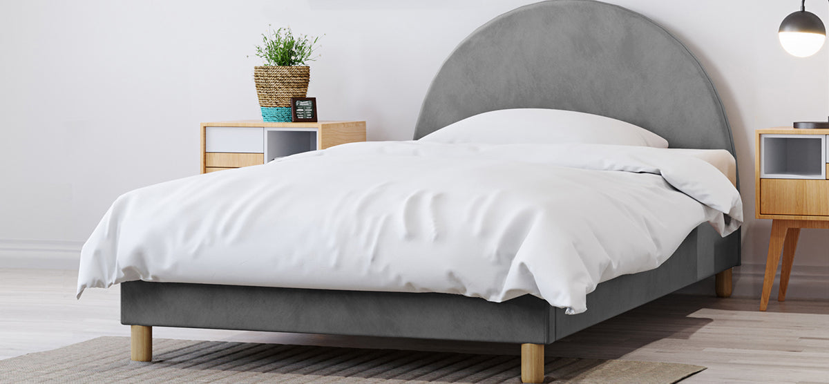 Minimalist bedroom with a low-profile upholstered bed featuring a grey headboard and white bedding, flanked by wooden nightstands with a plant, clock, and table lamp.