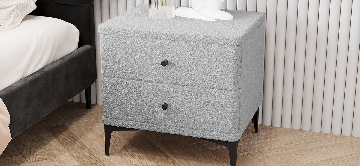 A light boucle-upholstered nightstand with black metal legs, topped with a glass vase of tall greenery and a white bust, stands on herringbone hardwood flooring.