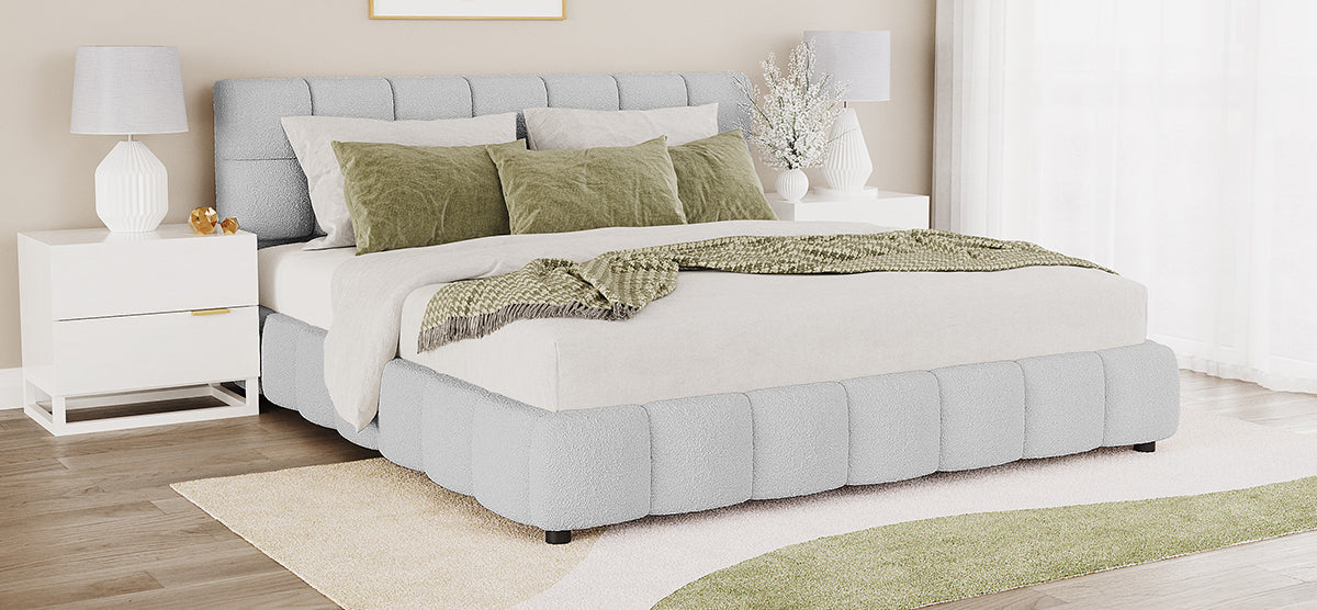 Contemporary bedroom with a low-profile pale gray boucle upholstered bed, tufted headboard, and neutral linens. Flanked by white nightstands with sculptural lamps. Green throw and pillows add color.