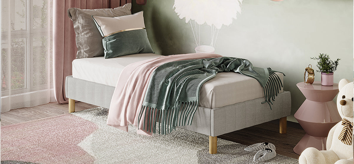 Cozy bedroom corner with a light-gray upholstered single bed, decorative pillows, throws, plush rug, pink side table with a vintage clock, and a cream teddy bear.