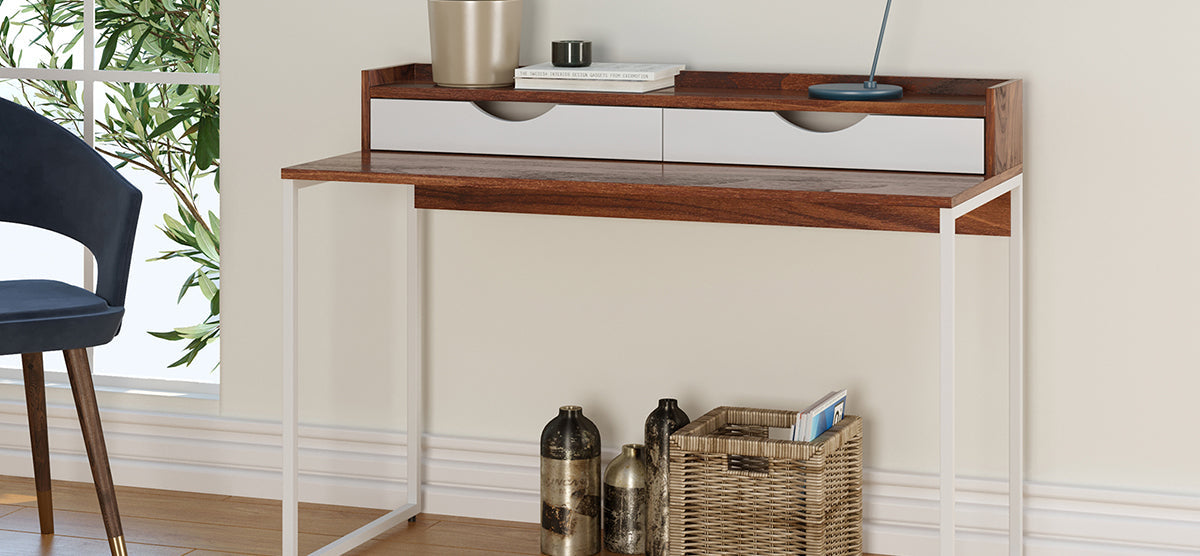 Modern writing desk with a warm wood-grain top, two white-front drawers, and slim white legs. Decor includes books, a planter, desk lamp, and nearby blue chair.