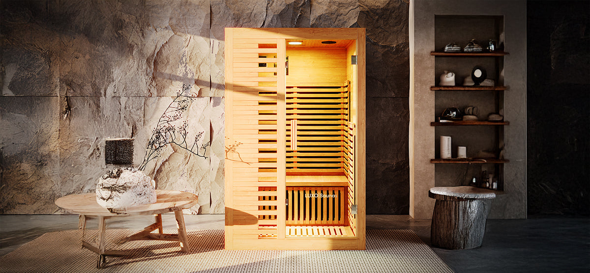Freestanding indoor sauna cabin made of light-colored wood, illuminated by an amber glow. Accompanied by a wooden table with a vase, shelving with towels, and a rustic stool.