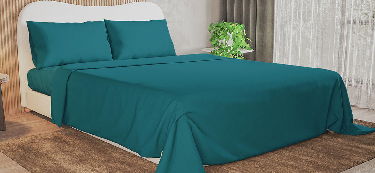 Neatly made bed with teal bedding and white headboard, beside a round table with a potted plant. Brown area rug and sheer curtains complete the cozy bedroom setting.