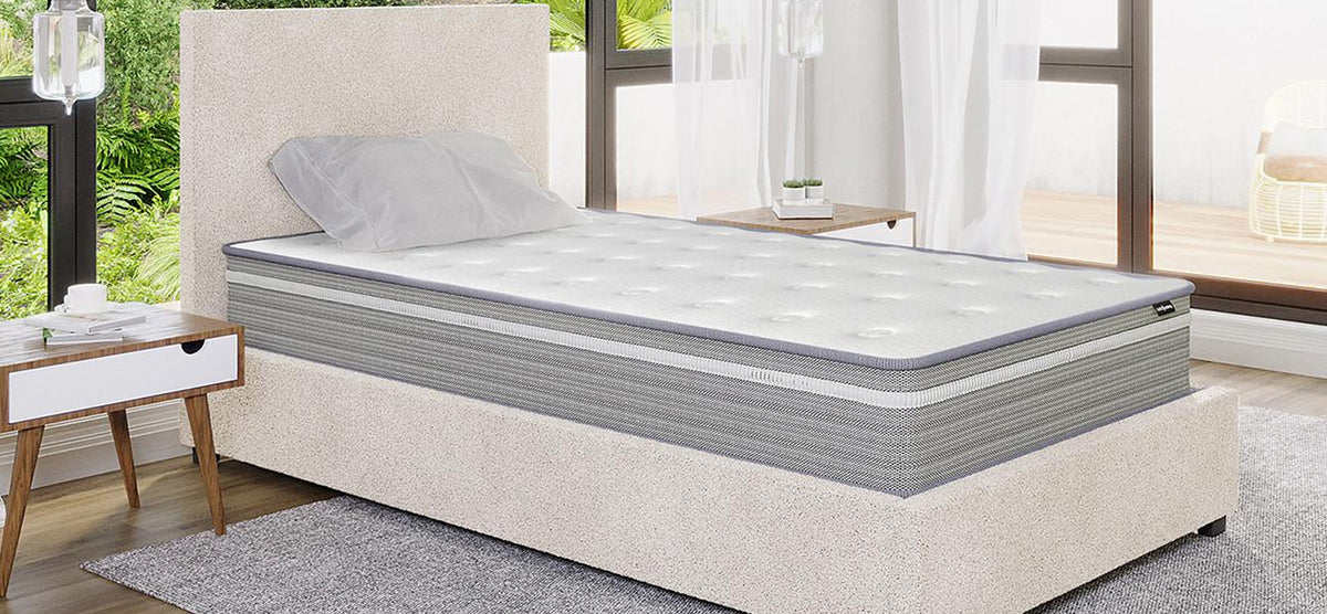 Minimalist bedroom with a light-beige upholstered platform bed, gray-and-white mattress, small wooden nightstand with coffee cup and book, neutral rug, and sheer-curtained windows overlooking greenery.