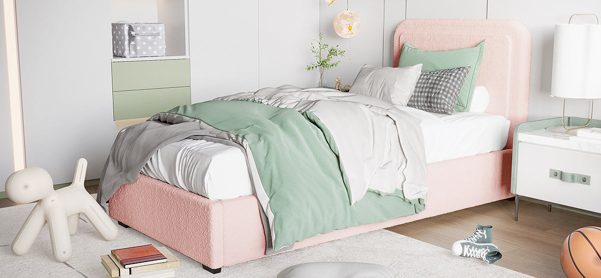 A modern bedroom corner with a pale pink upholstered bed, muted green and gray bedding, mint-green sneakers, a sculptural dog seat, and white nightstand with a gold-trim lamp.