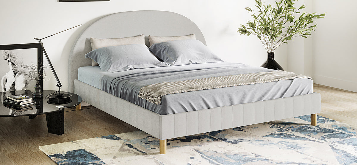 Minimalist modern bedroom featuring a light gray upholstered platform bed, black side table with lamp, books, horse sketch, abstract rug, and tall green plant in a black vase.