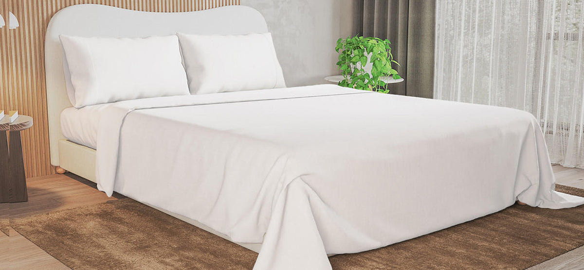 Modern bedroom with a neatly made bed in white linens and a light-wood headboard. Features include a potted plant and book on side tables, and sheer curtains.