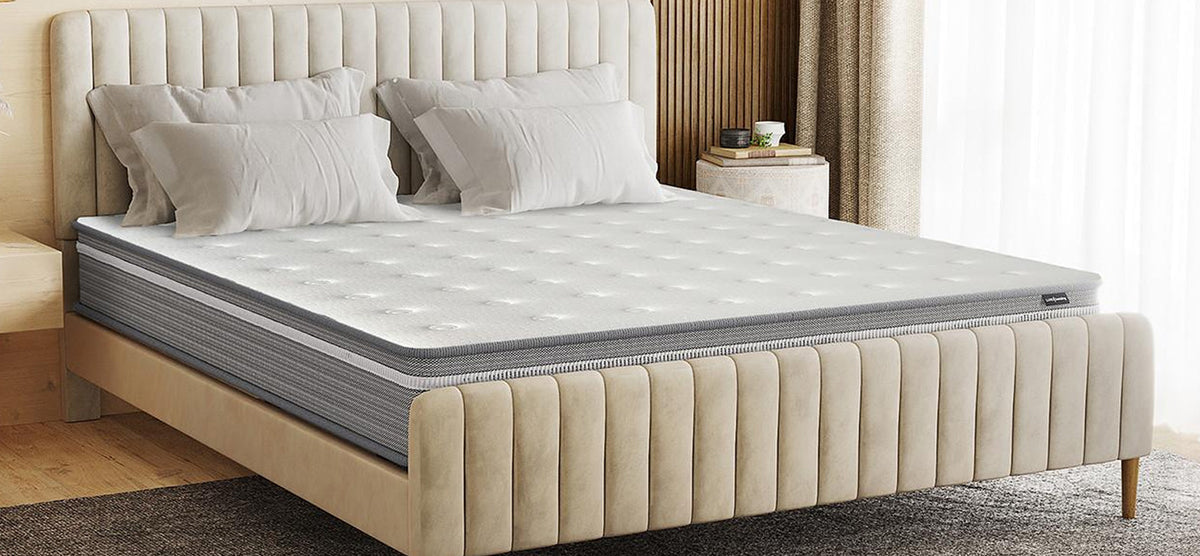 Neatly made bed with a beige velvet upholstered frame, channel-tufted headboard and footboard, gray quilted mattress, white pillows, wooden nightstand with books, plant, sheer curtains, and a textured rug.